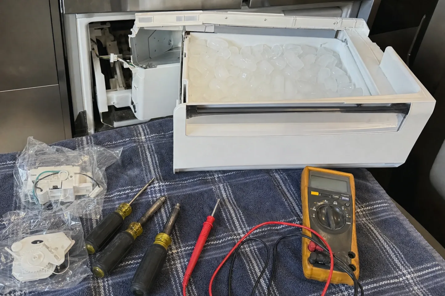 Ice drawer set on a work towel beside two bagged replacement parts, screwdrivers and a multimeter